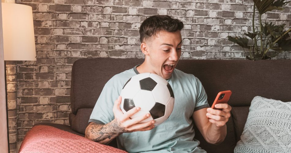 Young boy soccer fan watching a football game on mobile phone sitting on a comfortable sofa in living room