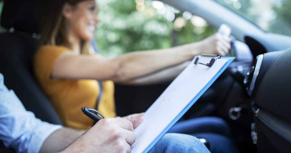 Close up view of driving instructor holding checklist while in background female student steering and driving car.